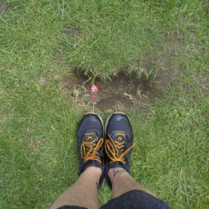 photographie de deux pieds chaussés de souliers avec lacets oranges. Les pieds sont devant un trou de terre dans l'herbe avec une petite fleur